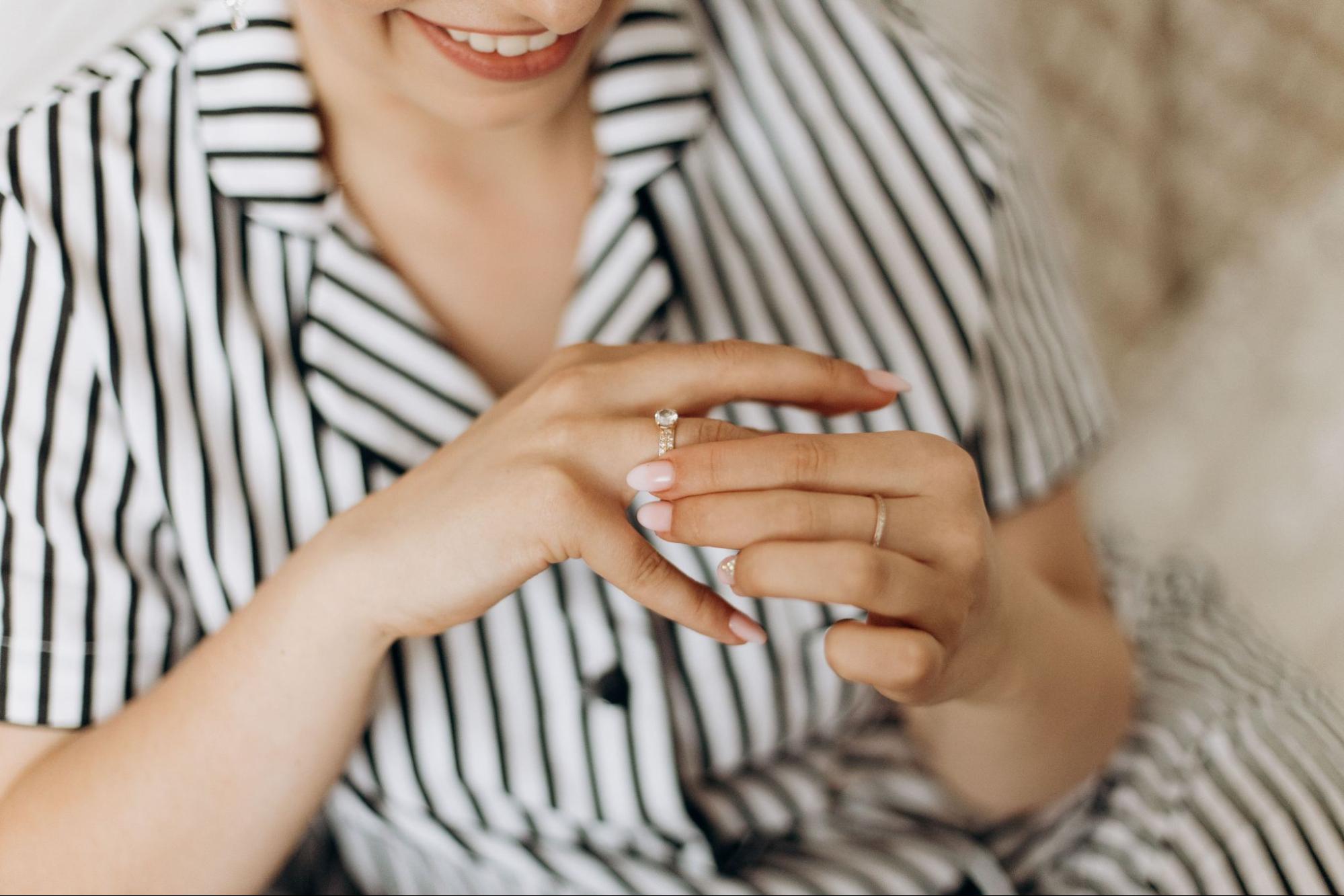 A smiling woman wearing a striped shirt admires her diamond engagement ring while touching it with her other hand. A smiling woman wearing a striped shirt admires her diamond engagement ring while touching it with her other hand.
