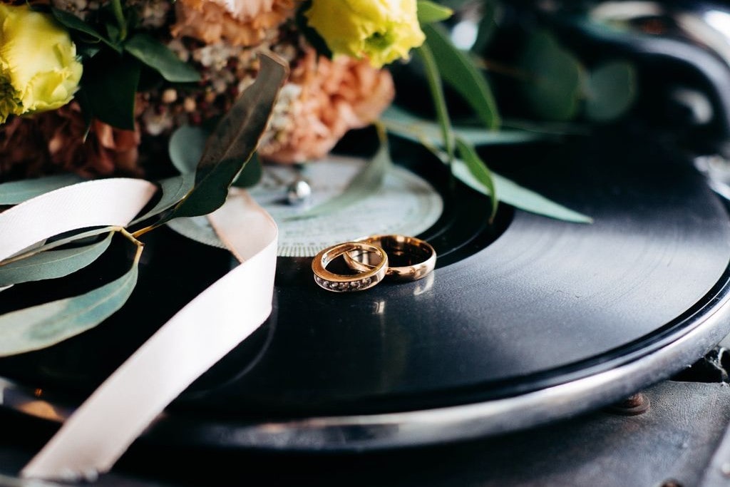 A pair of gold wedding bands resting on a vinyl record, surrounded by flowers and a satin ribbon. A pair of gold wedding bands resting on a vinyl record, surrounded by flowers and a satin ribbon.