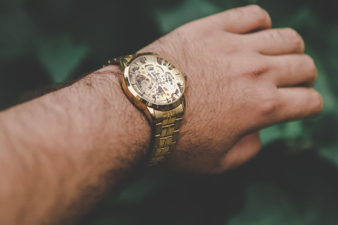 A man’s wrist displaying a vintage-style, gold timepiece while on a walk in a garden