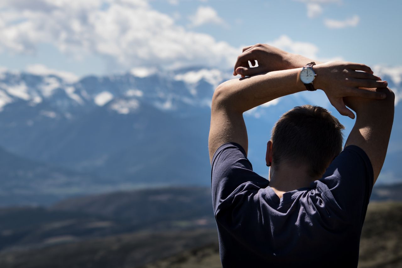 A man stretching with his arms over his head. You can see the fine watch on his wrist and mountains in the background