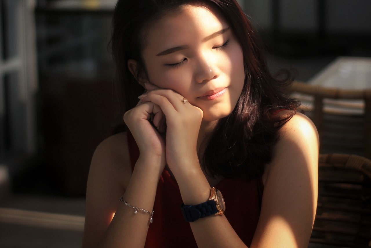 A sunbeam crosses a woman’s face and she closes her eyes as she rests her hands against her face as she sits at a restaurant table. She has a luxury watch, charm bracelet, and gold fashion ring on