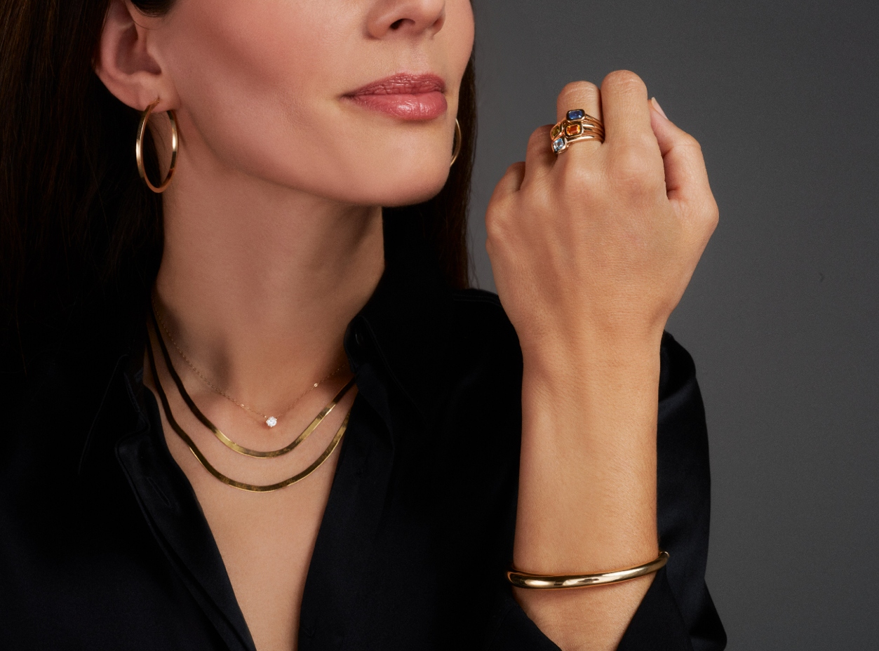 A sophisticated woman with gold hoop earrings, two gold necklaces, a gold bangle, and multiple gemstone gold rings worn in an artful stack.