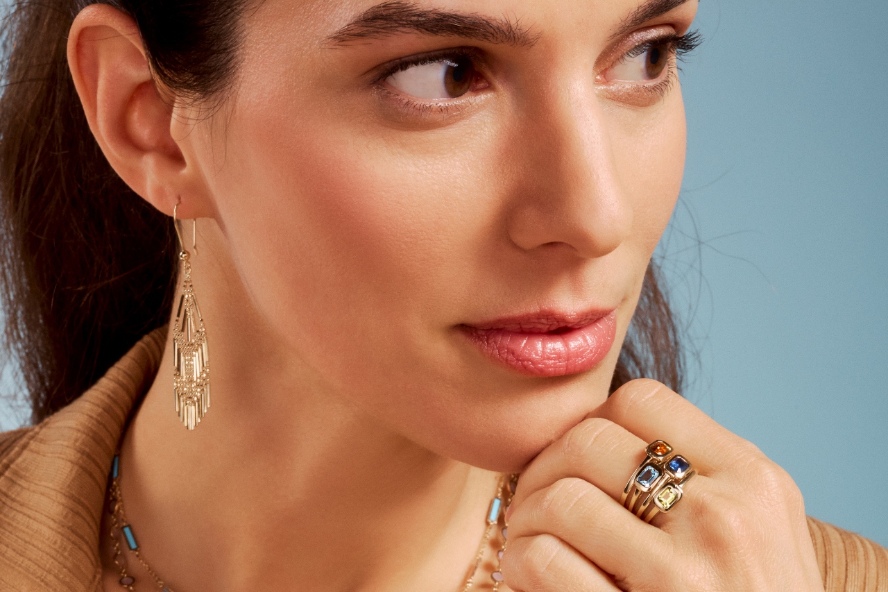 A woman wearing gold chandelier earrings, a gemstone gold necklace, and multiple gemstone gold rings.