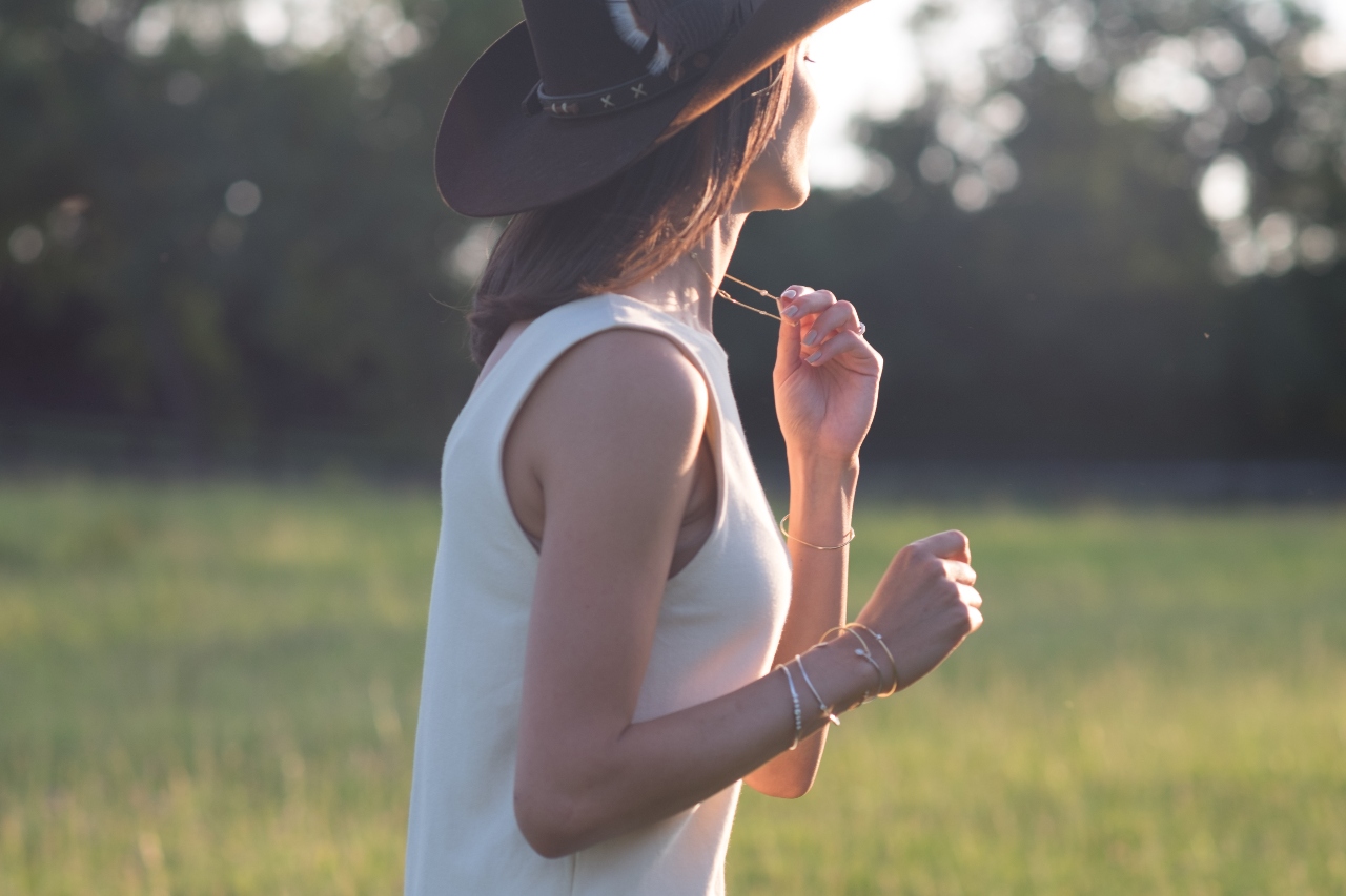 A woman standing in a field with a white tank top and brown cowboy hat with matching yellow gold bracelets along her wrists and a gold necklace around her neck that she’s holding onto with one hand.