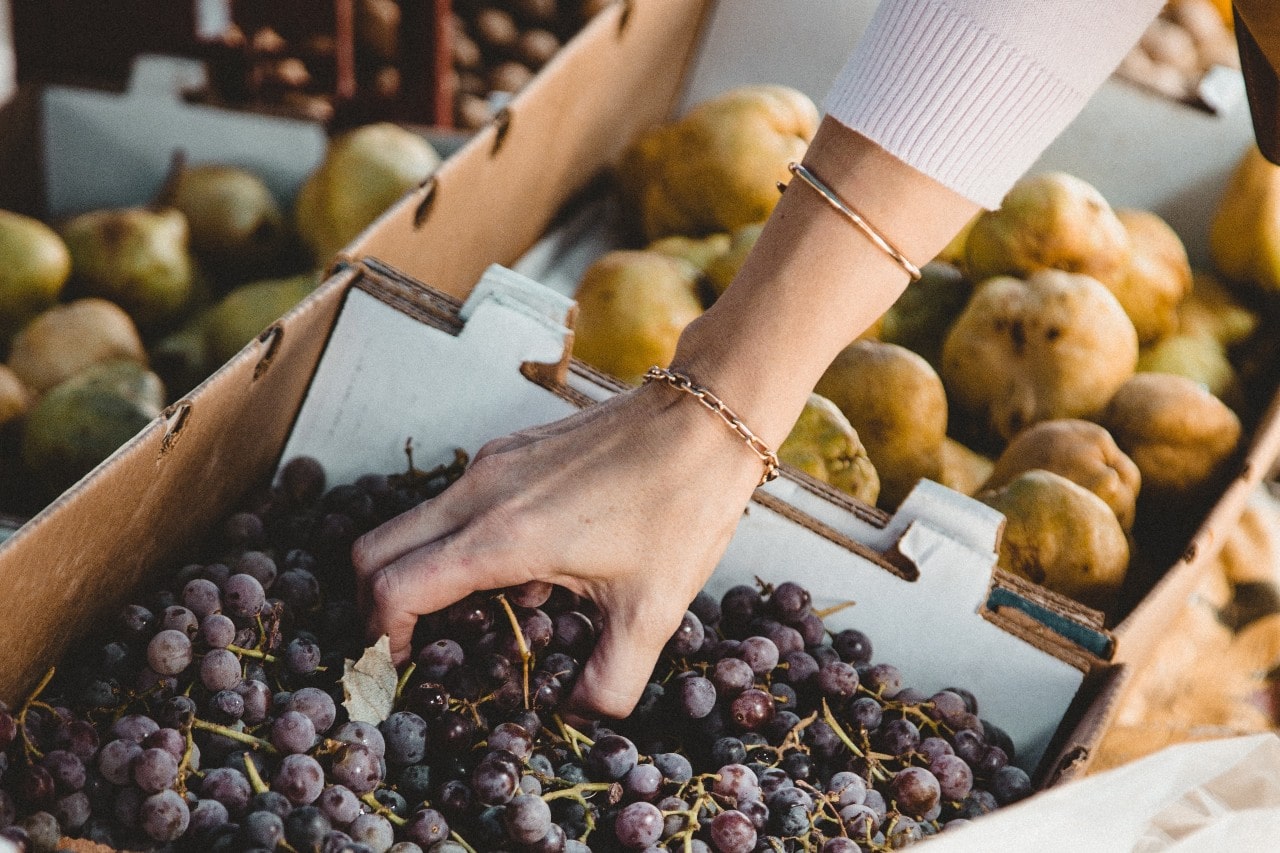 A close-up of a woman’s hand selecting grapes at a market, adorned two different stacked bracelets.