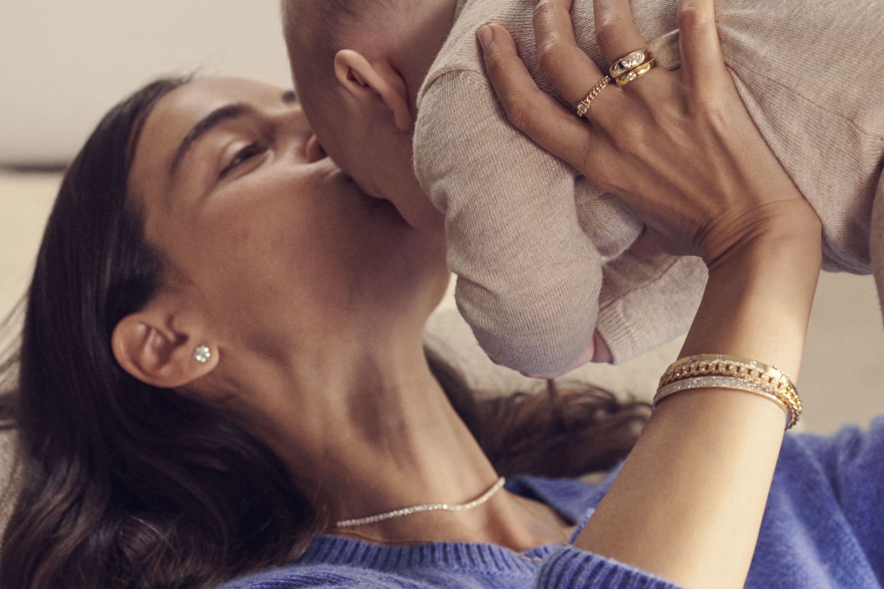 A woman holding and giving a kiss to a baby while wearing gold and diamond bracelets stacked together with a matching diamond collar necklace and round diamond stud earrings.