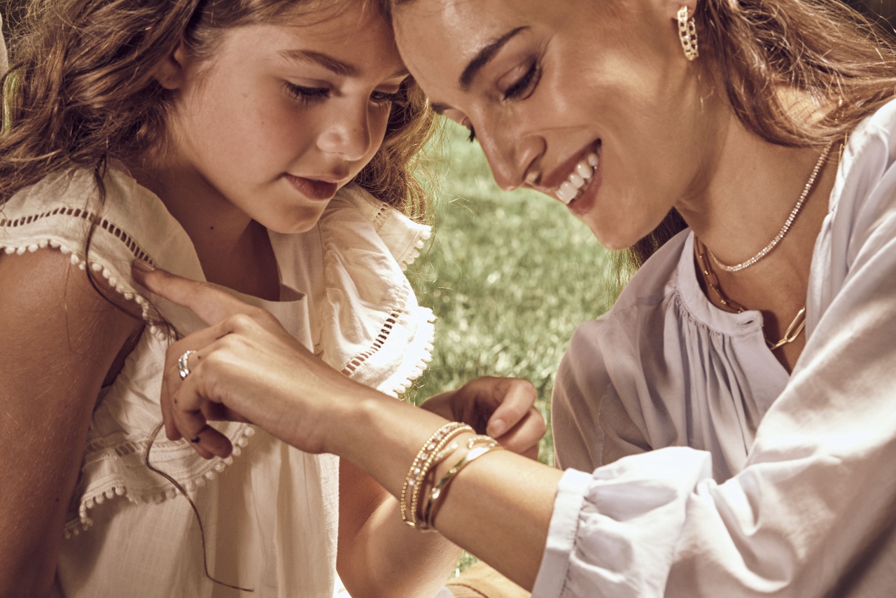 A young daughter admiring her mother’s gold and diamond bracelet stack while they picnic on the grass. 